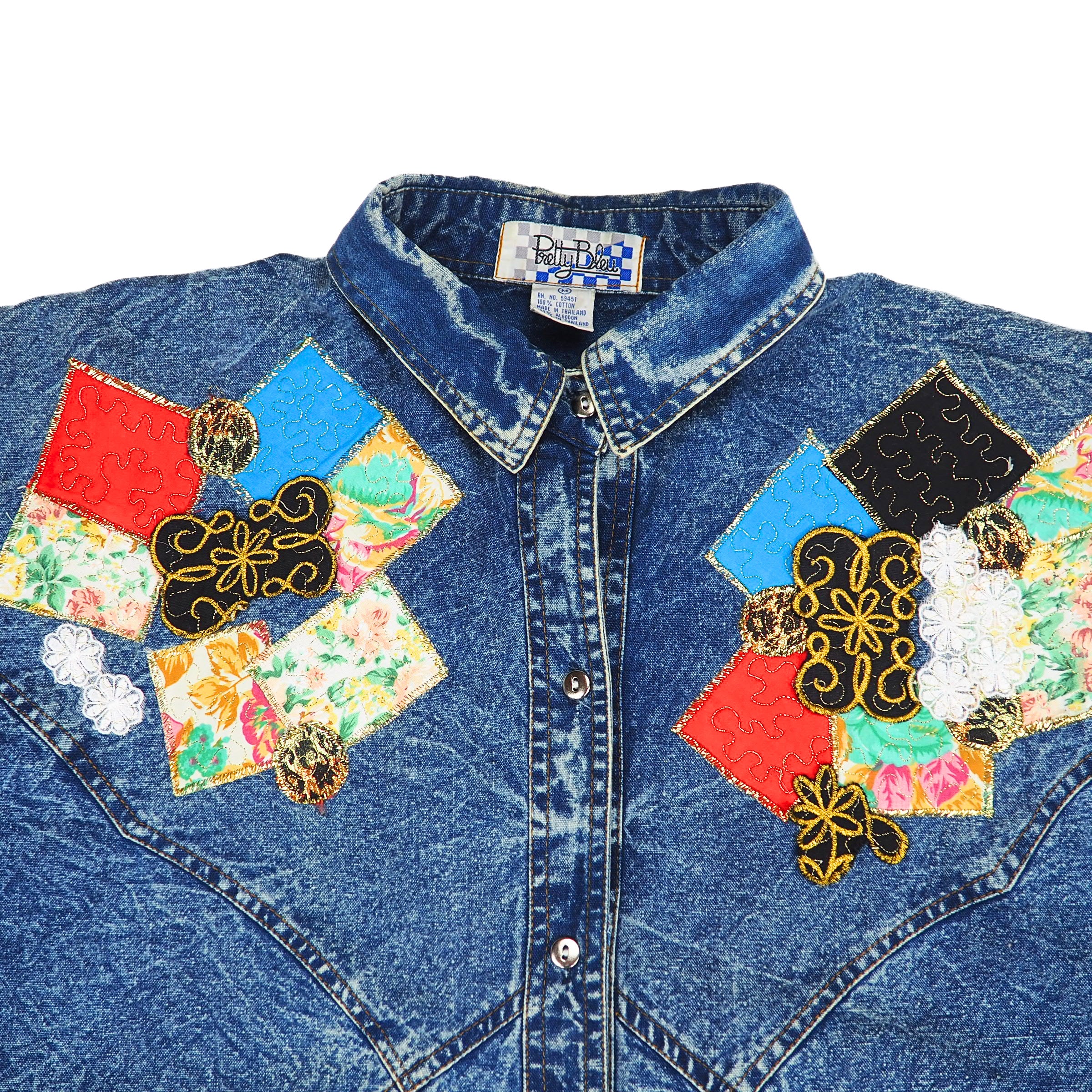 Detail of collar and chest of a vintage acid wash blue denim shirt with colorful patchwork and embroidery on a white background
