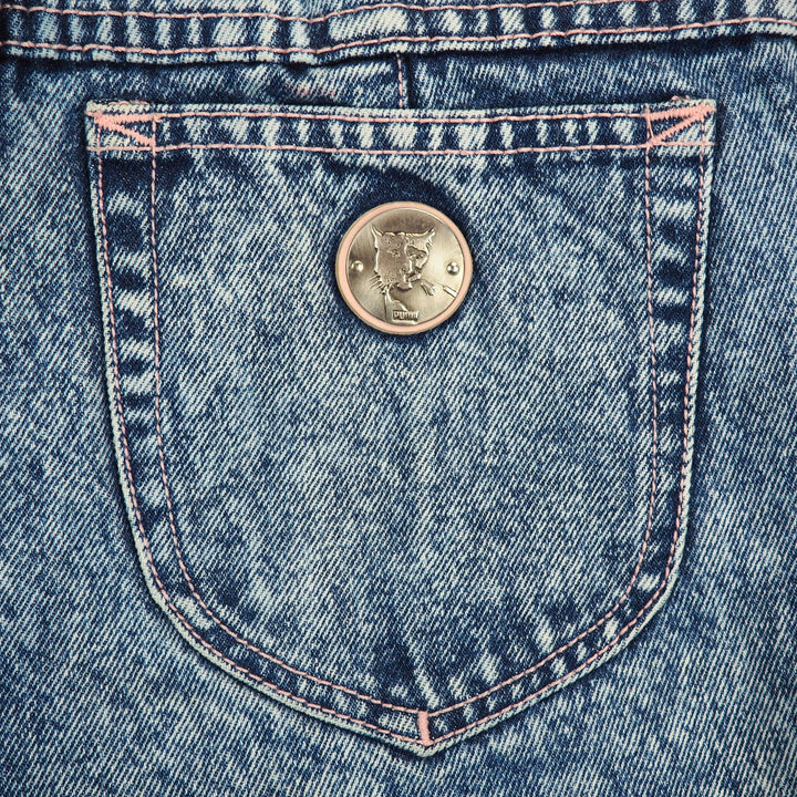 Close-up of a round coin like silver Puma cat logo on the back pocket of a pair of acid wash denim jeans.
