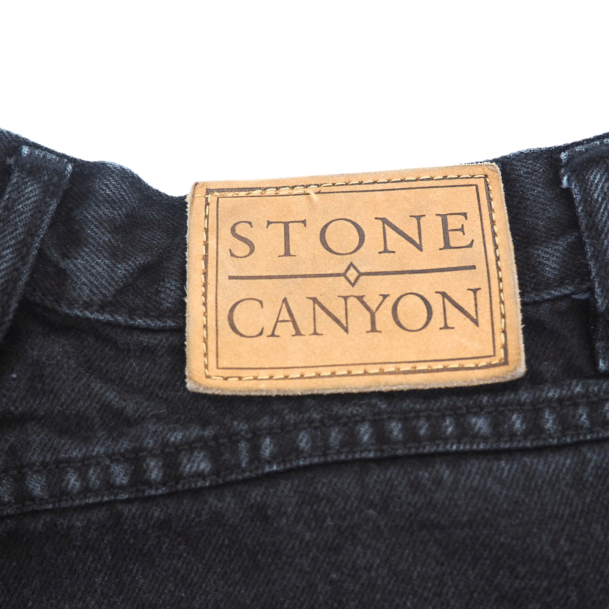 Close-up of a brown leather 'Stone Canyon' jarcon label on the waistband of a pair of black denim jeans.