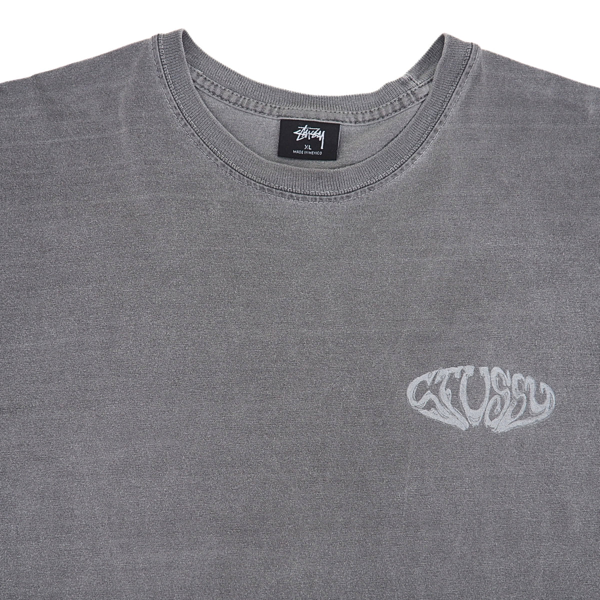 A close-up of a gray Stüssy crewneck t-shirt featuring a white Stüssy logo on the left chest, with the brand's black neck label visible inside the collar.
