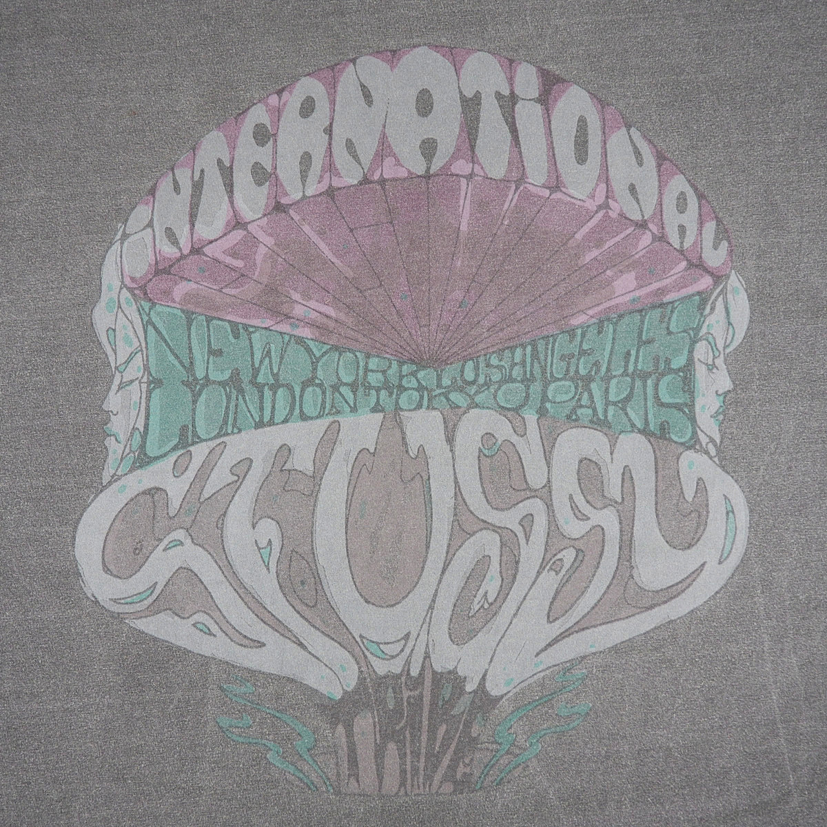 A gray t-shirt featuring a retro psychedelic graphic print. The design shows a stylized mushroom cap with faces on the sides, containing the words 'INTERNATIONAL' in white at the top and 'STUSSY' in white at the bottom, with a teal band across the middle listing cities including New York, Los Angeles, London, Toronto, and Paris.
