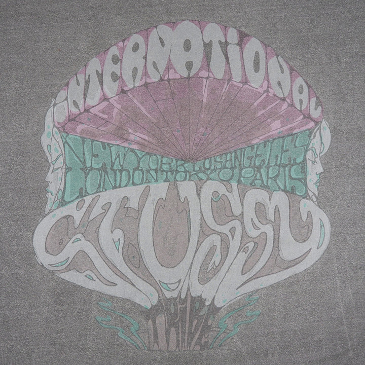 A gray t-shirt featuring a retro psychedelic graphic print. The design shows a stylized mushroom cap with faces on the sides, containing the words 'INTERNATIONAL' in white at the top and 'STUSSY' in white at the bottom, with a teal band across the middle listing cities including New York, Los Angeles, London, Toronto, and Paris.