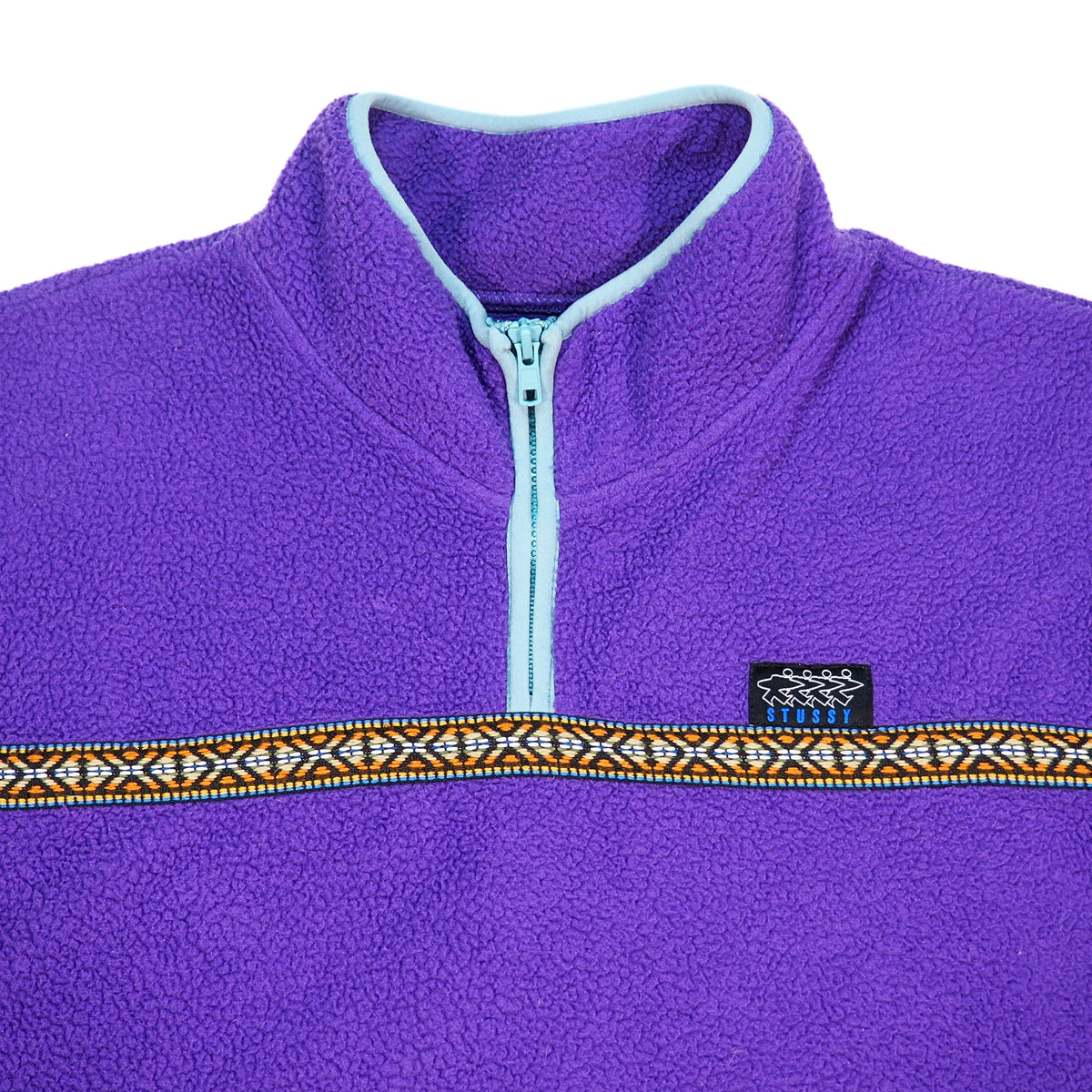 Close up of the neck area of a purple fleece quarter-zip pullover jacket with a light blue zipper, light blue trim, and a decorative horizontal patterned stripe across the chest. A small black Stussy logo is visible on the chest.
