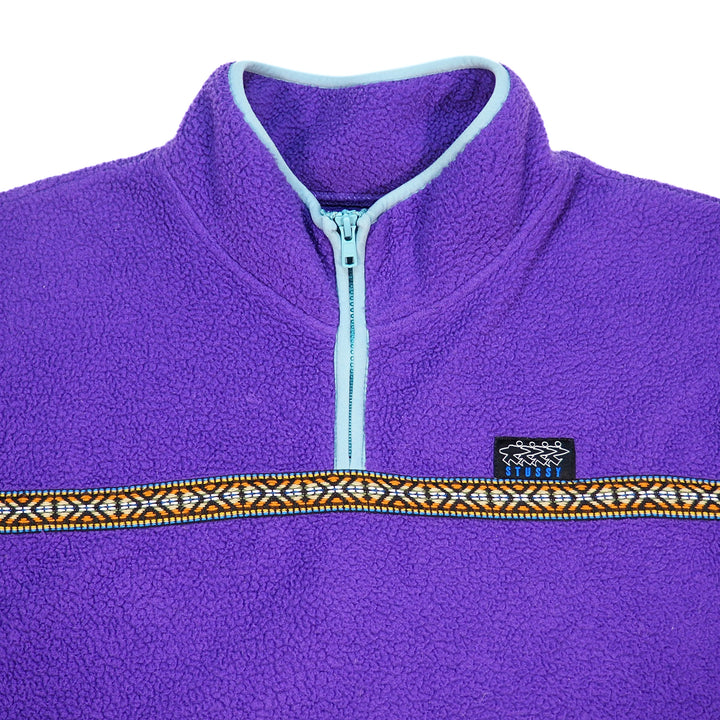 Close up of the neck area of a purple fleece quarter-zip pullover jacket with a light blue zipper, light blue trim, and a decorative horizontal patterned stripe across the chest. A small black Stussy logo is visible on the chest.