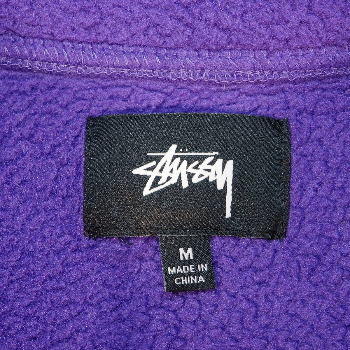 Close up of a black Stussy inner size label with size M and Made in China on a purple fleece garment.