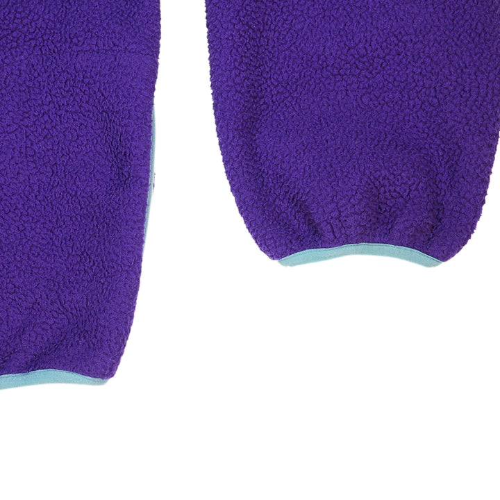 Close up of the sleeve cuff and hem of a purple fleece sweater with blue trim on a white background.