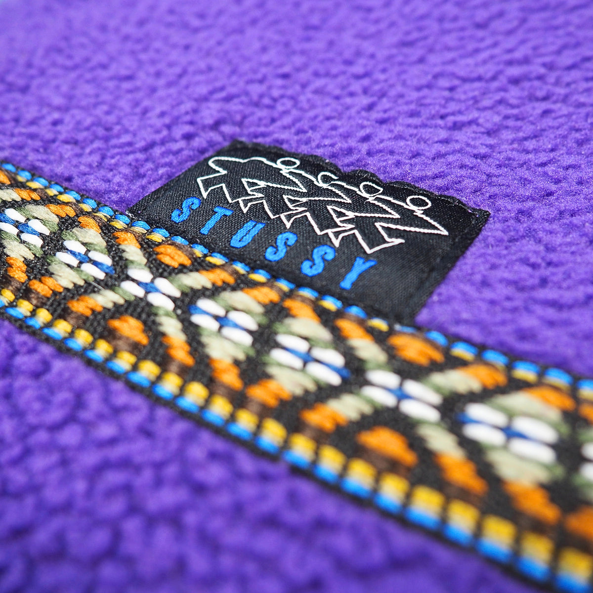 Close up of a black Stussy label and embroidered multi-color tape on a pruple fleece sweater.