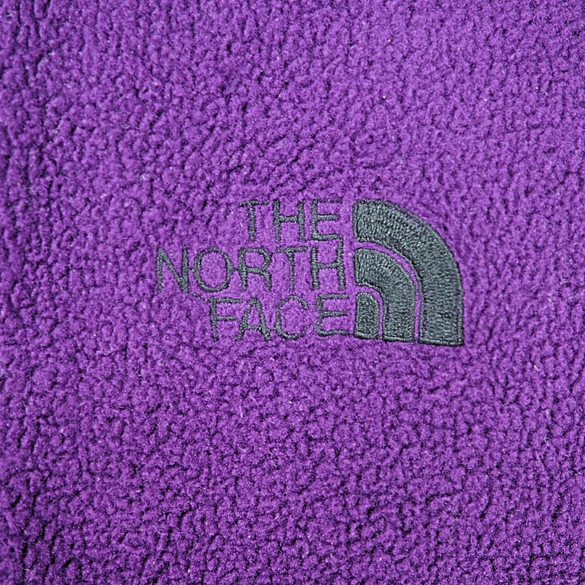 Close up of an embroidered black The North Face logo on purple fleece fabric.
