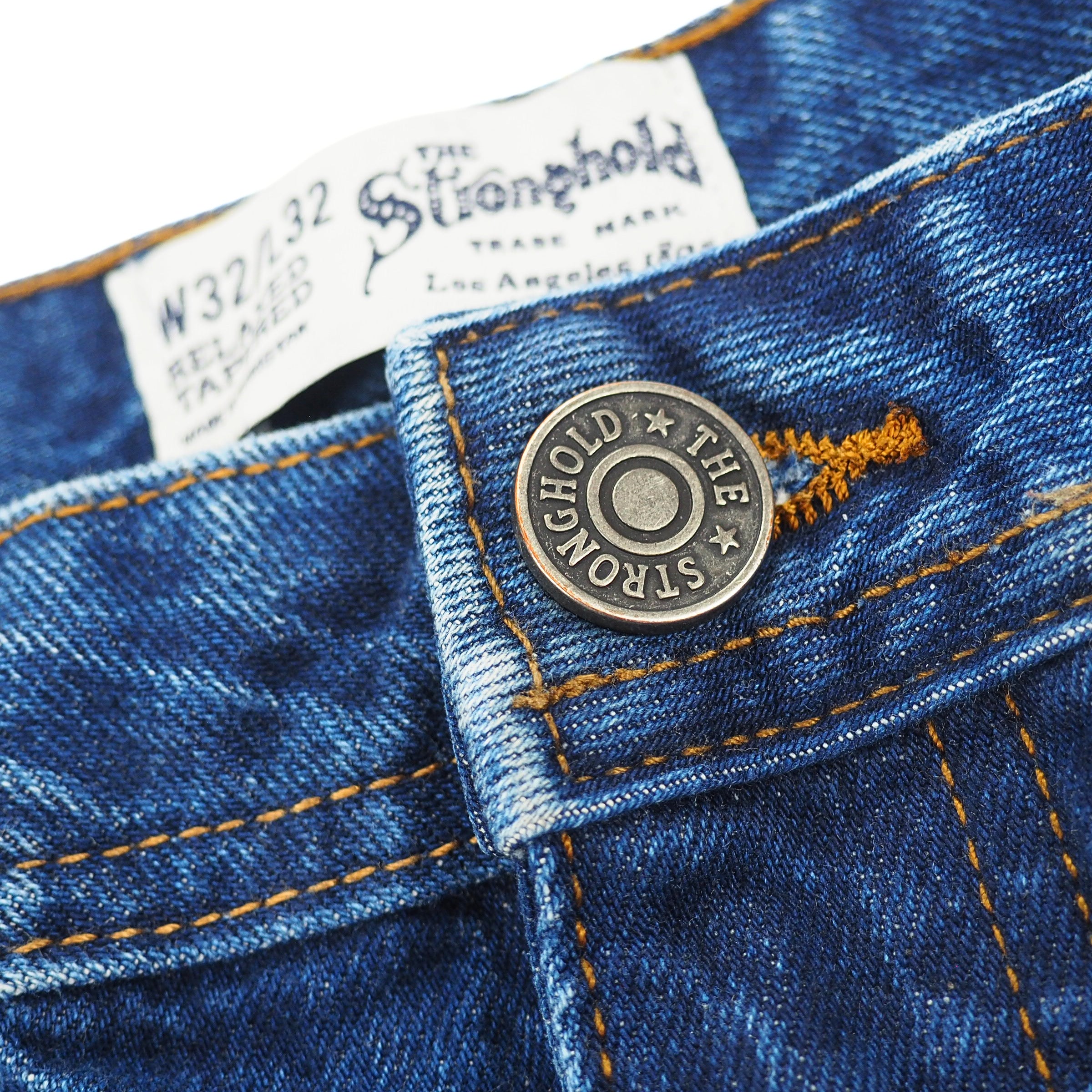 Close-up of blue denim jeans with The Stronghold brand label and stamped metal button.