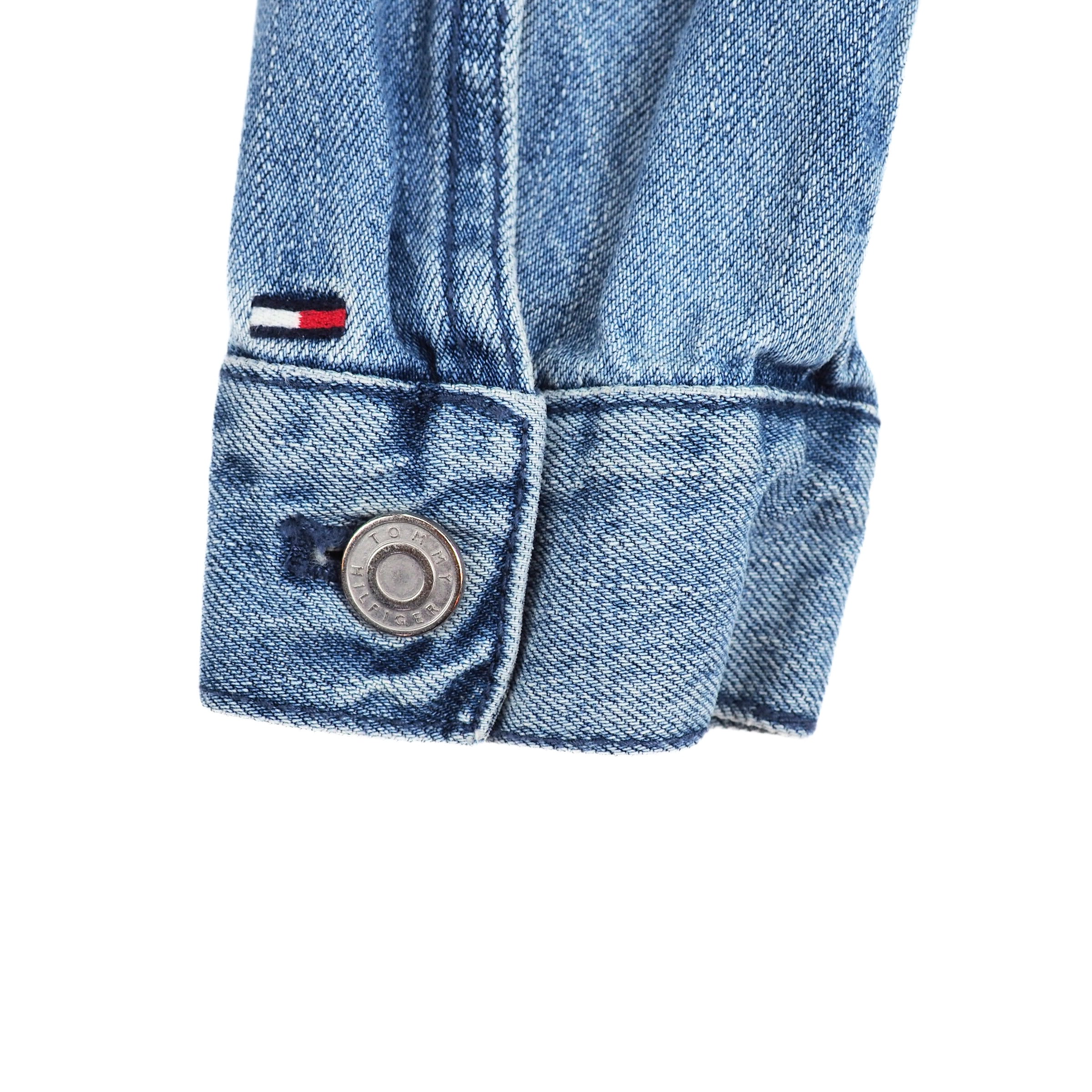 Close-up of a blue denim jacket sleeve cuff with a Tommy Hilfiger flag brand logo and stamped button on a white background