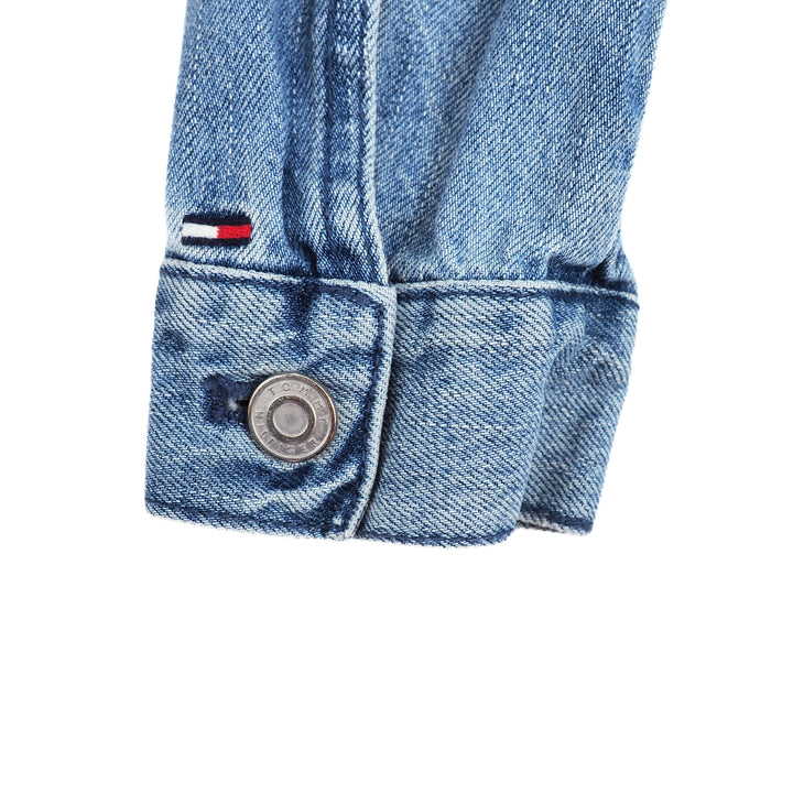 Close-up of a blue denim jacket sleeve cuff with a Tommy Hilfiger flag brand logo and stamped button on a white background