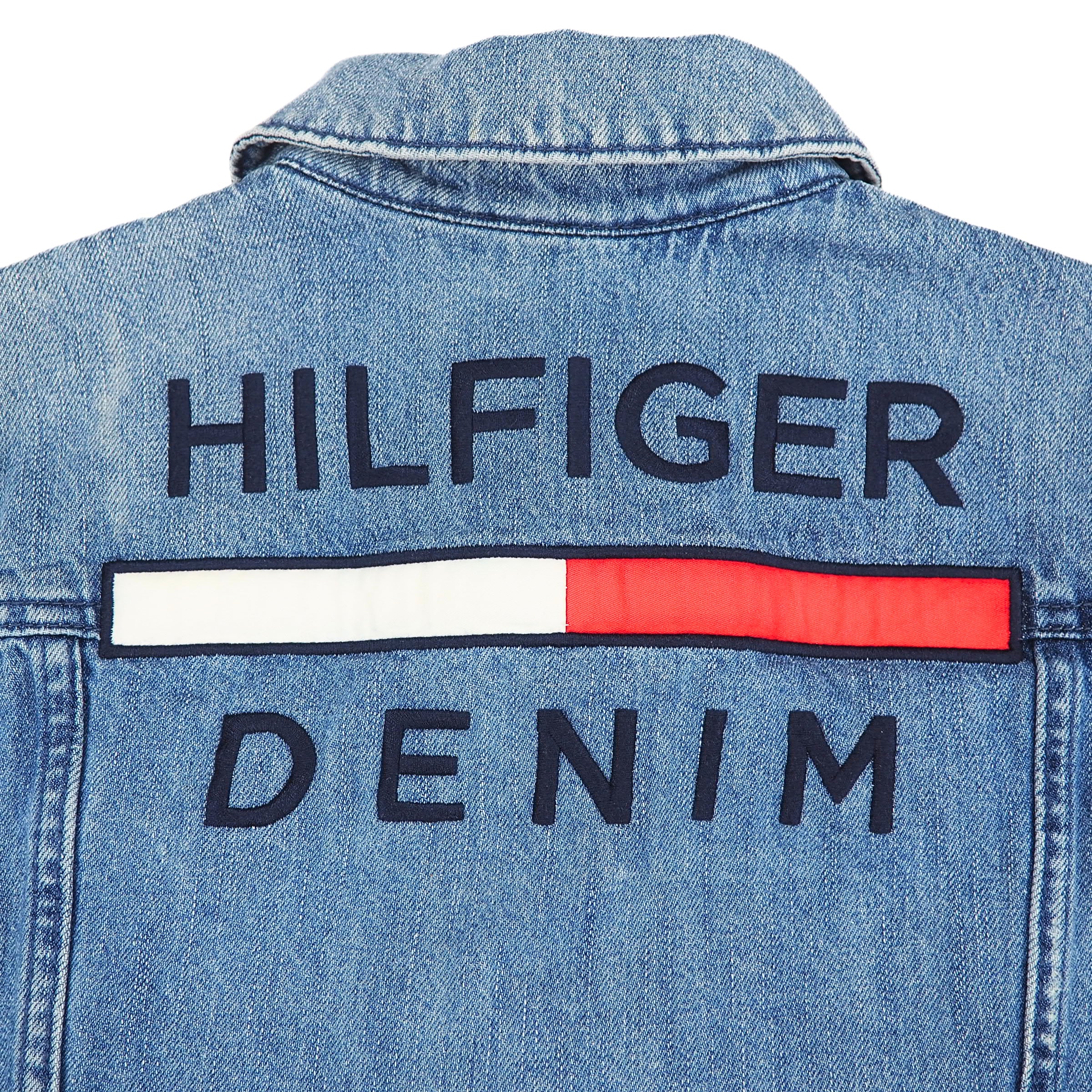 Close-up detail of a light blue denim jacket collar and shoulder with embroidered 'Hilfiger Denim' branding on a white background