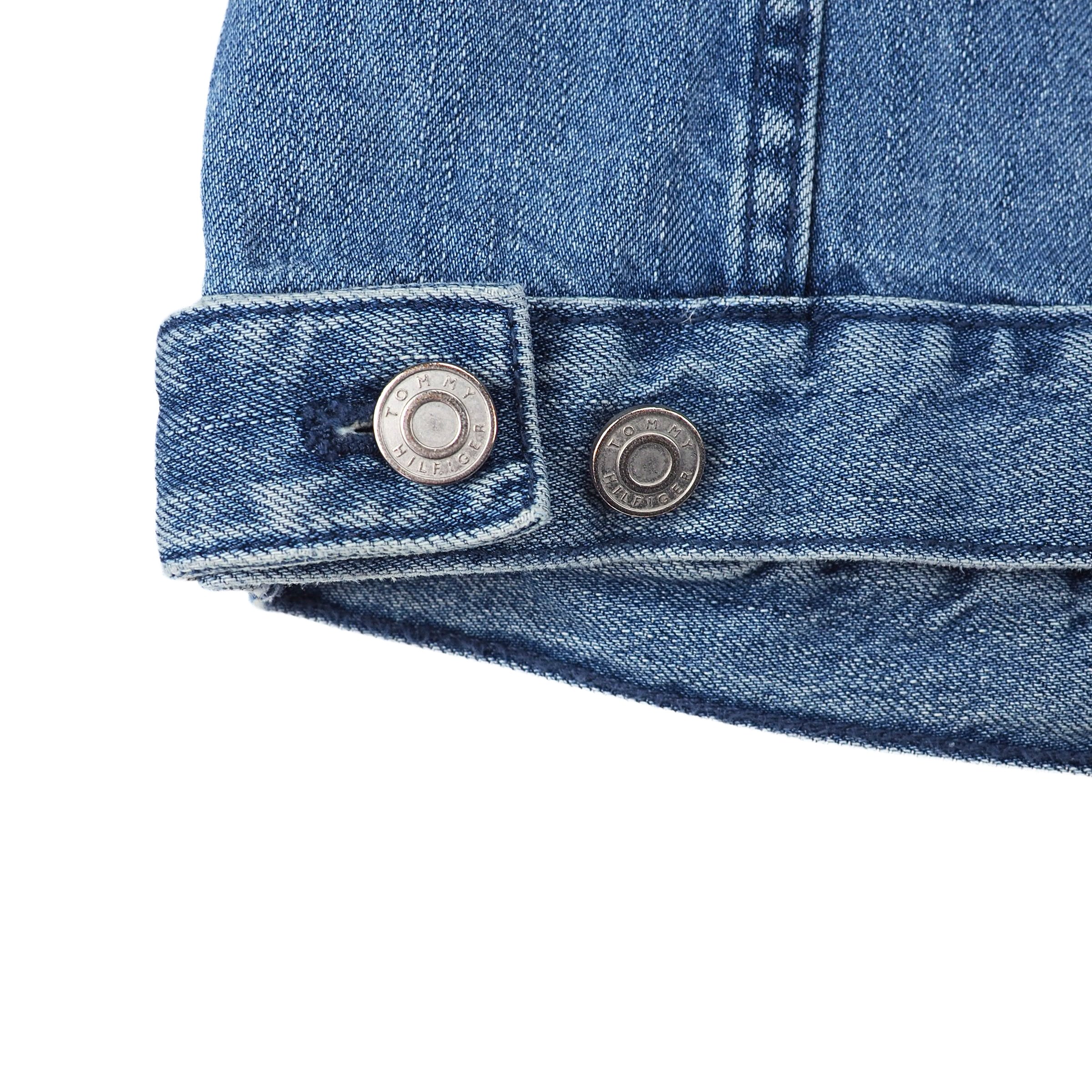 Close-up of a blue denim jacket hem with stamped 'Tommy Hilfiger' button details on a white background