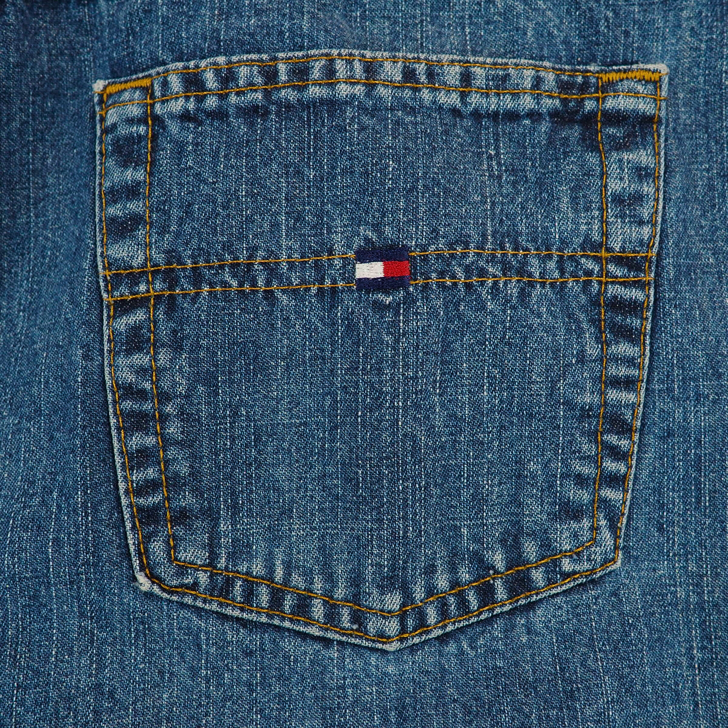 Close-up of a blue denim jean pocket with a Tommy Hilfiger flag logo and orange stitching.