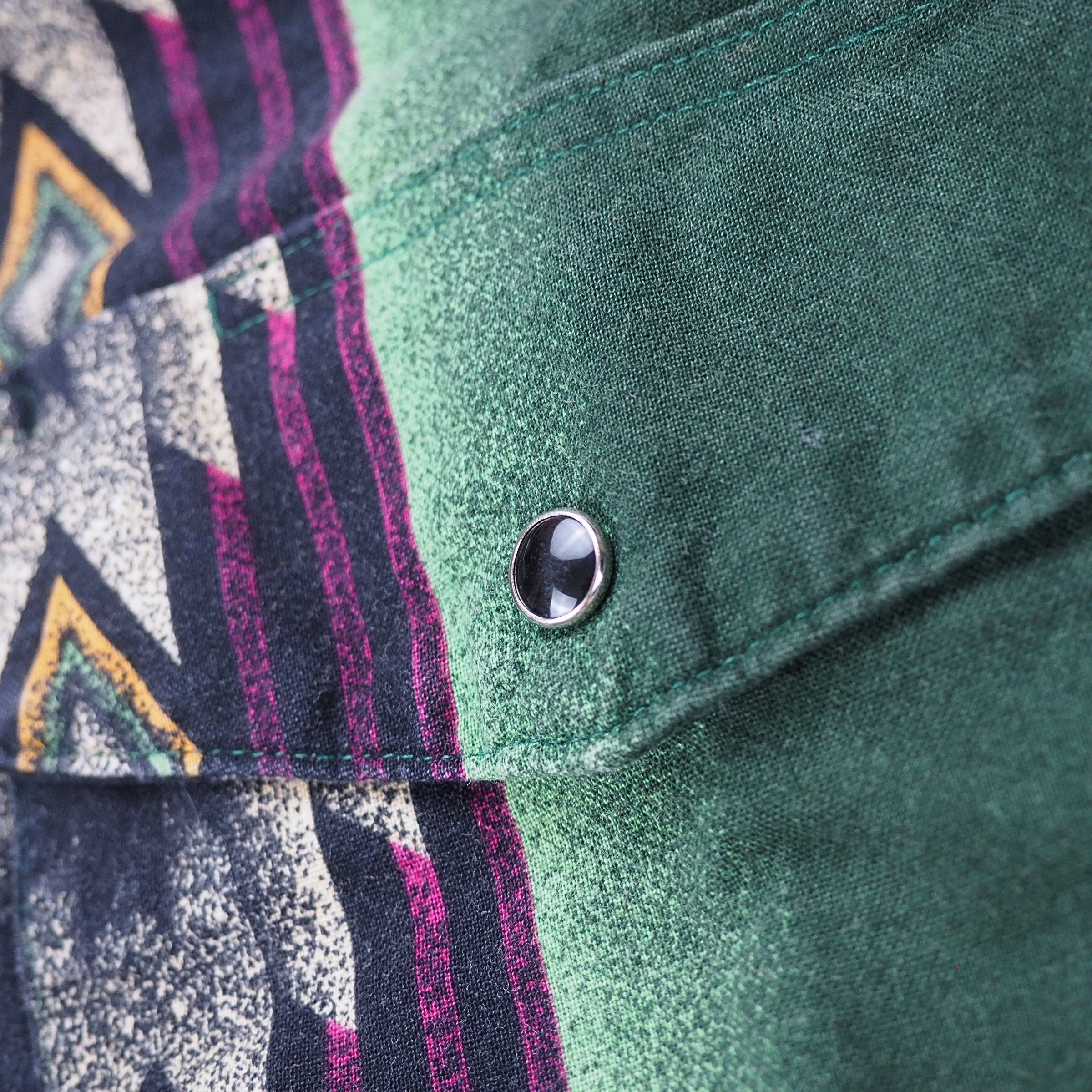 Close up of the pocket flap and onyx press stud button on a shirt with green, purple and orange pattern.