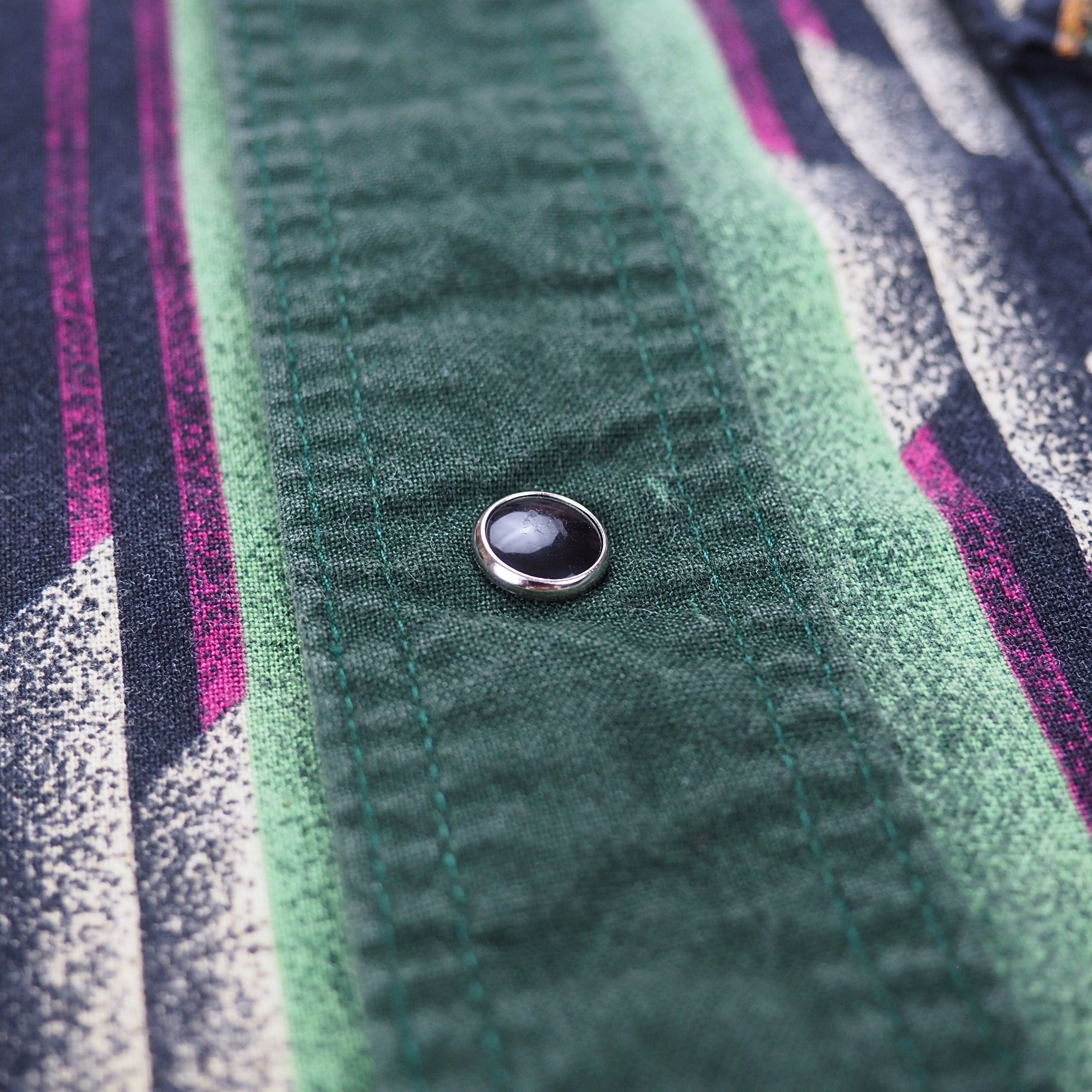 Close up of an onyx press stud on the placket of a green shirt with purple and white details.